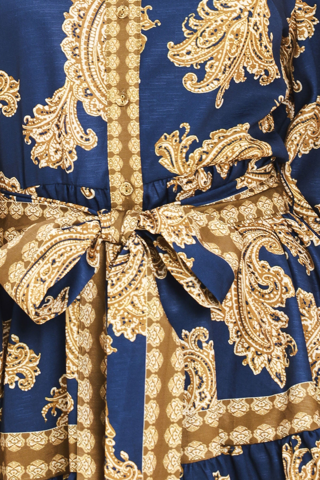 A woman modeling a long-sleeve mini dress with a blue and gold paisley pattern, featuring a button-up front, tie waist, and flared skirt | Flying Tomato