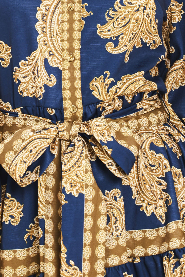 A woman modeling a long-sleeve mini dress with a blue and gold paisley pattern, featuring a button-up front, tie waist, and flared skirt | Flying Tomato
