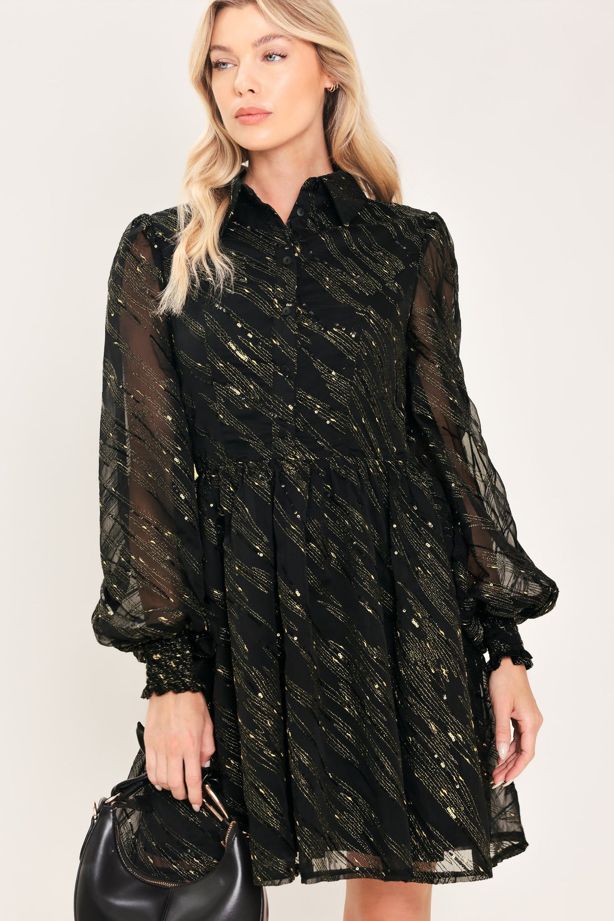 A model wearing a black sheer mini dress with gold glittery diagonal stripes, featuring long balloon sleeves and a flared skirt | Flying Tomato