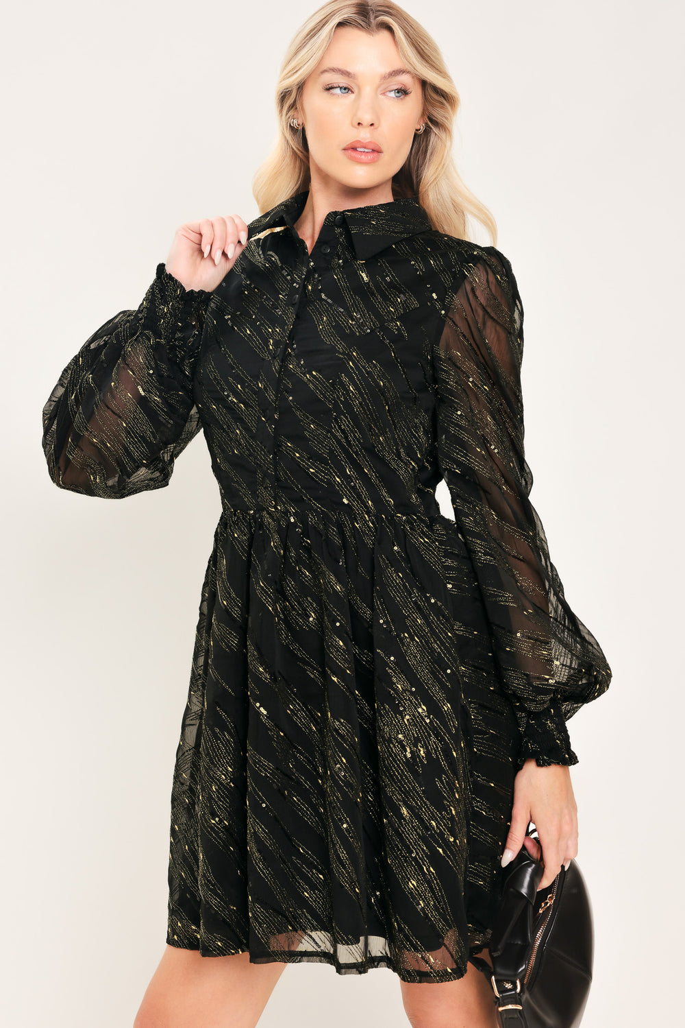 A model wearing a black sheer mini dress with gold glittery diagonal stripes, featuring long balloon sleeves and a flared skirt | Flying Tomato