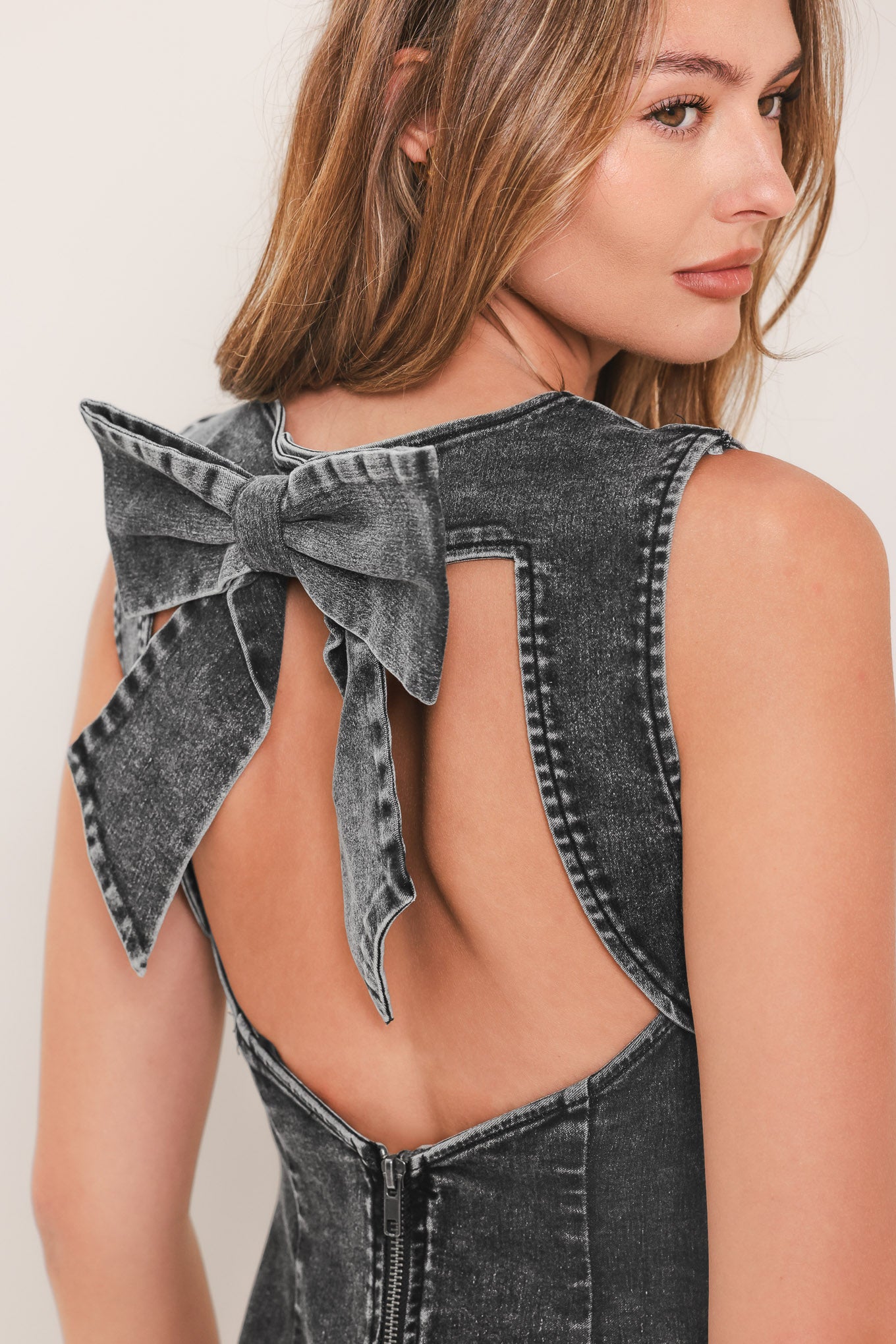 Close-up back view of a woman wearing a sleeveless dark gray denim dress with a large bow detail at the open back, against a plain light background.