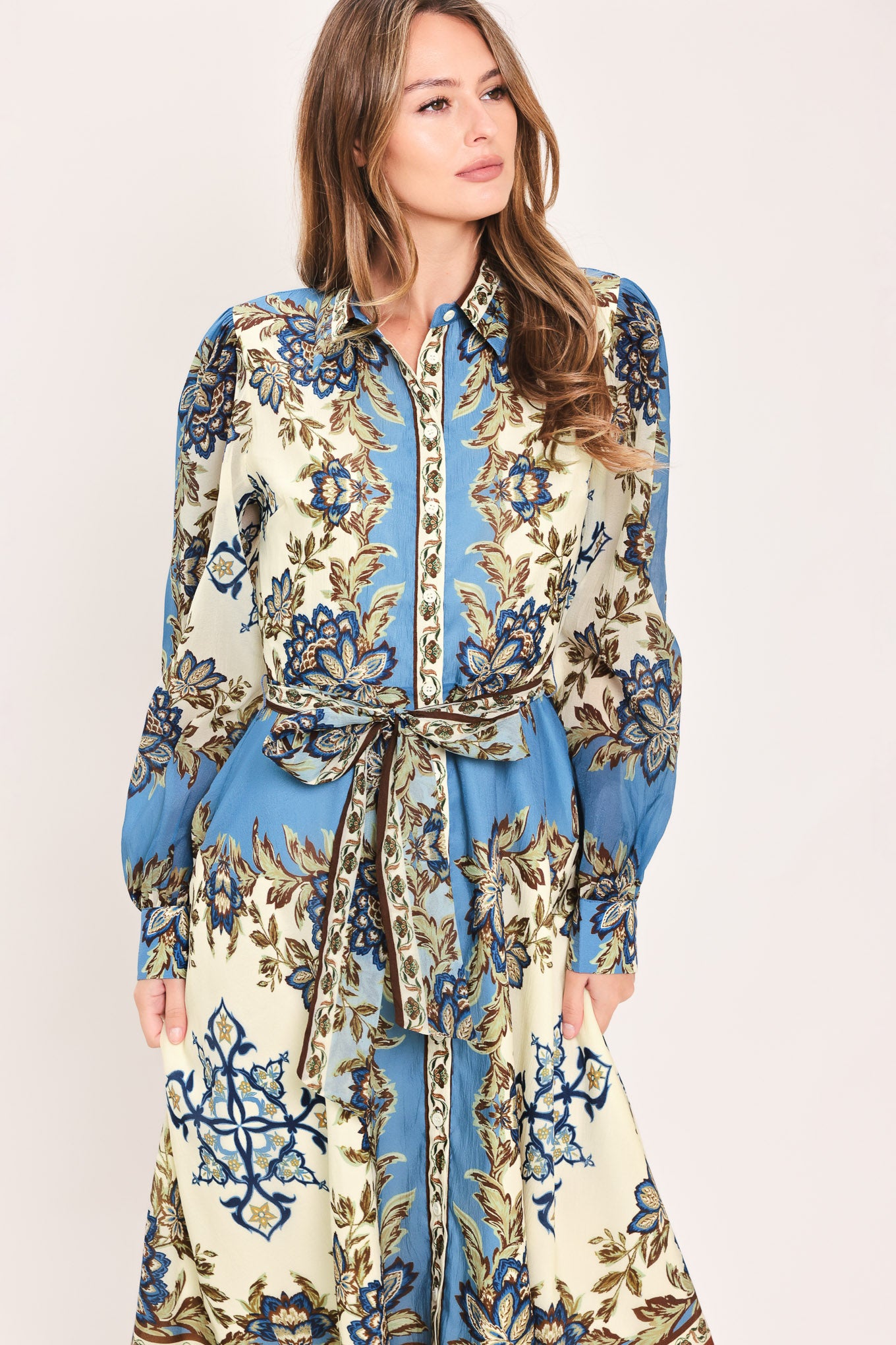 A woman wearing a long-sleeved midi dress with a blue, beige, and brown floral pattern, featuring a tie waist belt | Flying Tomato