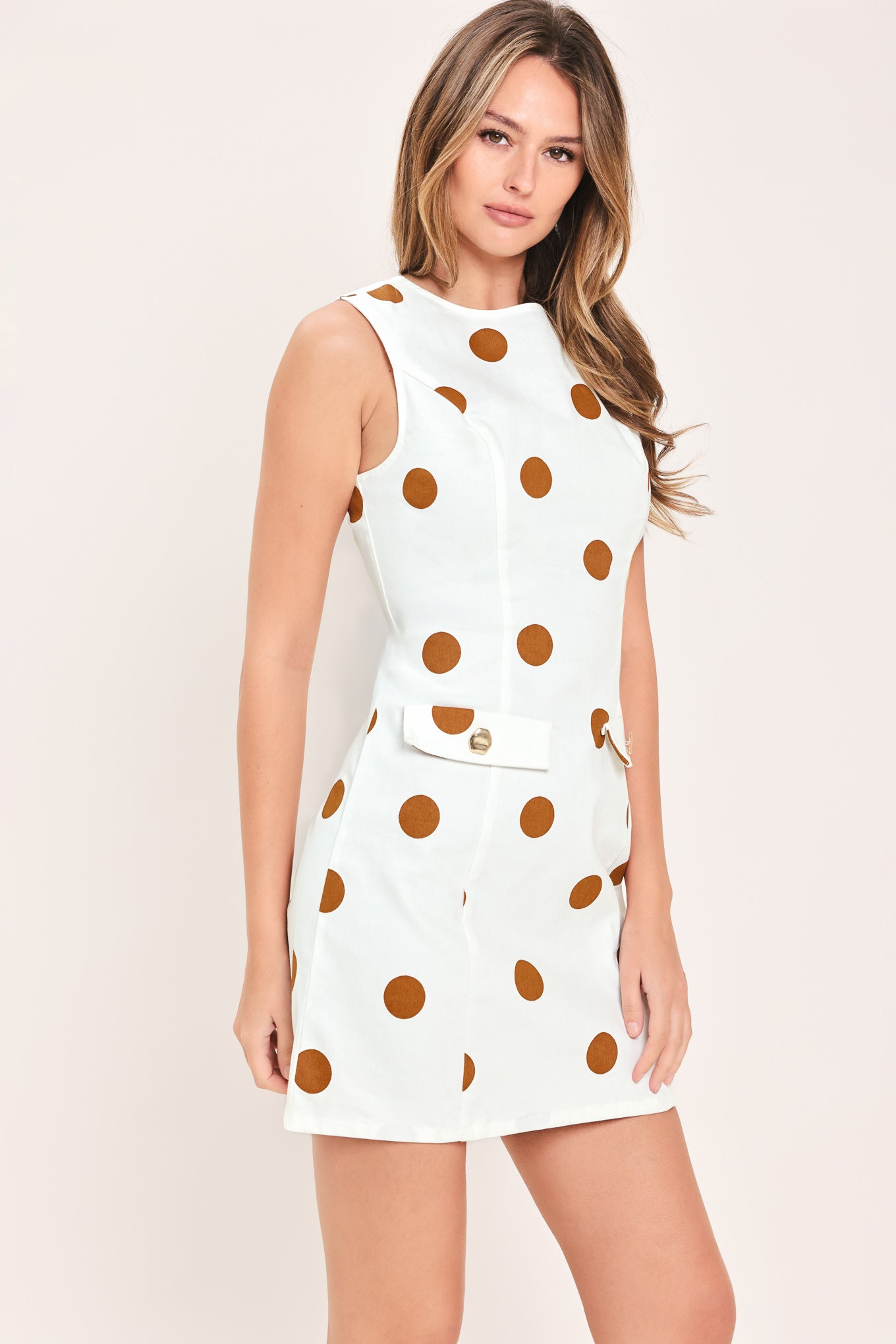 A woman wearing a sleeveless white mini dress with large brown polka dots and a small front flap pocket with a gold-tone button | Flying Tomato