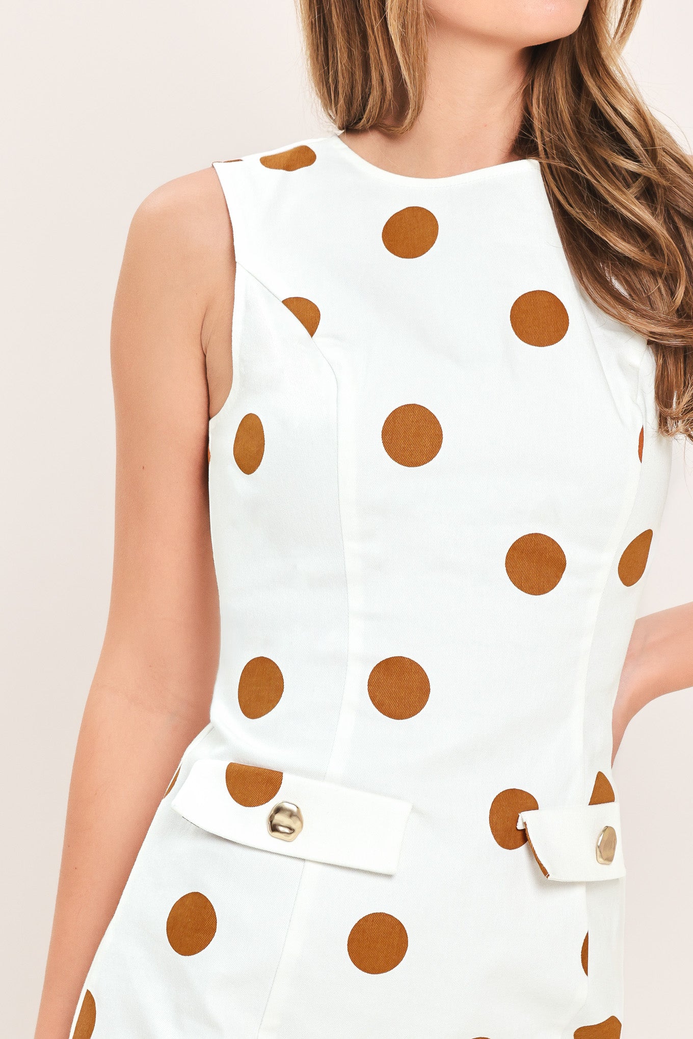 A woman wearing a sleeveless white mini dress with large brown polka dots and a small front flap pocket with a gold-tone button | Flying Tomato