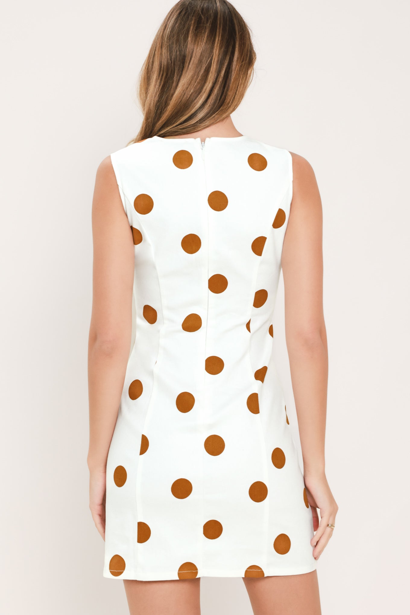 A woman wearing a sleeveless white mini dress with large brown polka dots and a small front flap pocket with a gold-tone button | Flying Tomato