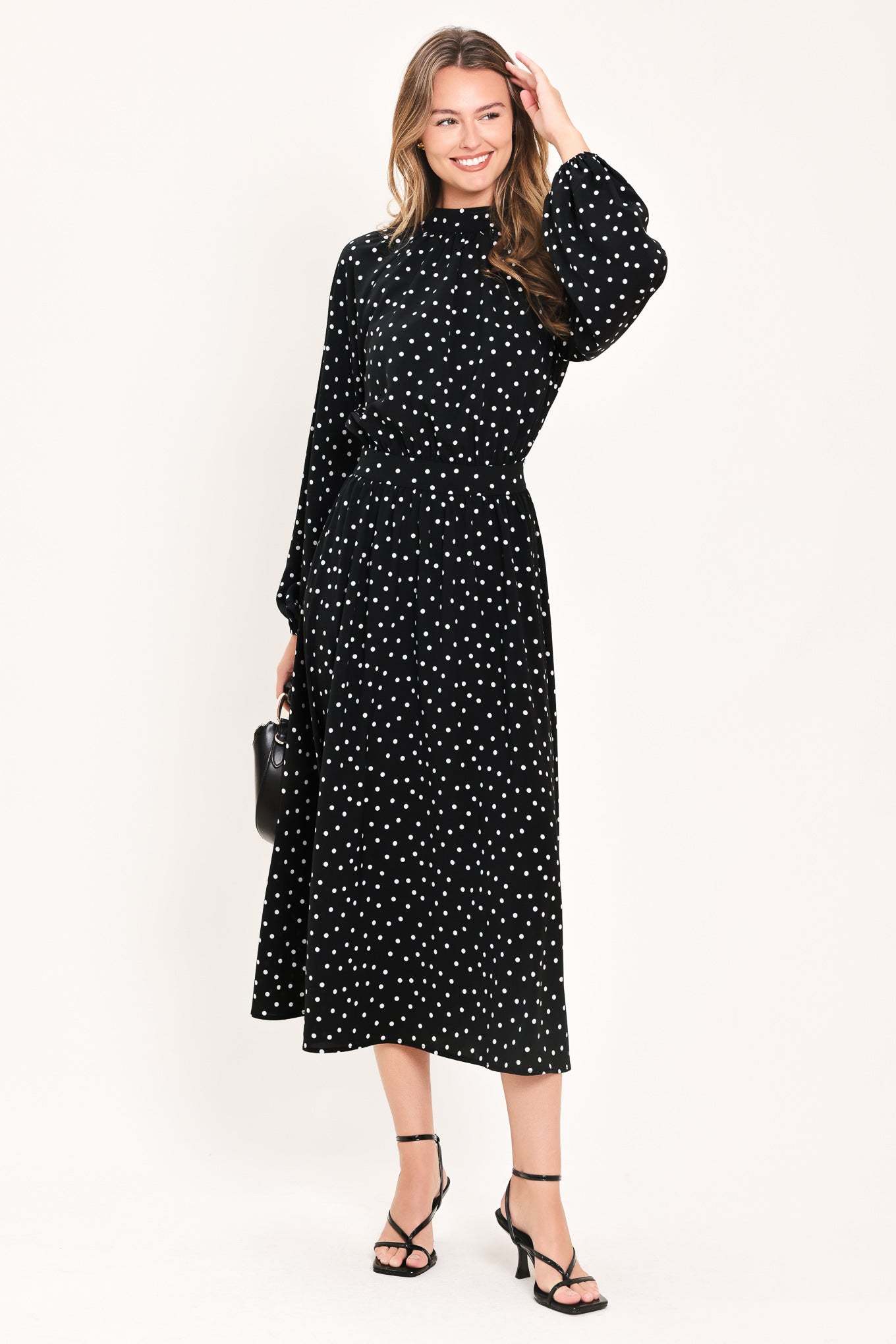 A woman modeling a black midi dress with a white polka dot pattern, featuring long balloon sleeves and a high mock neck | Flying Tomato