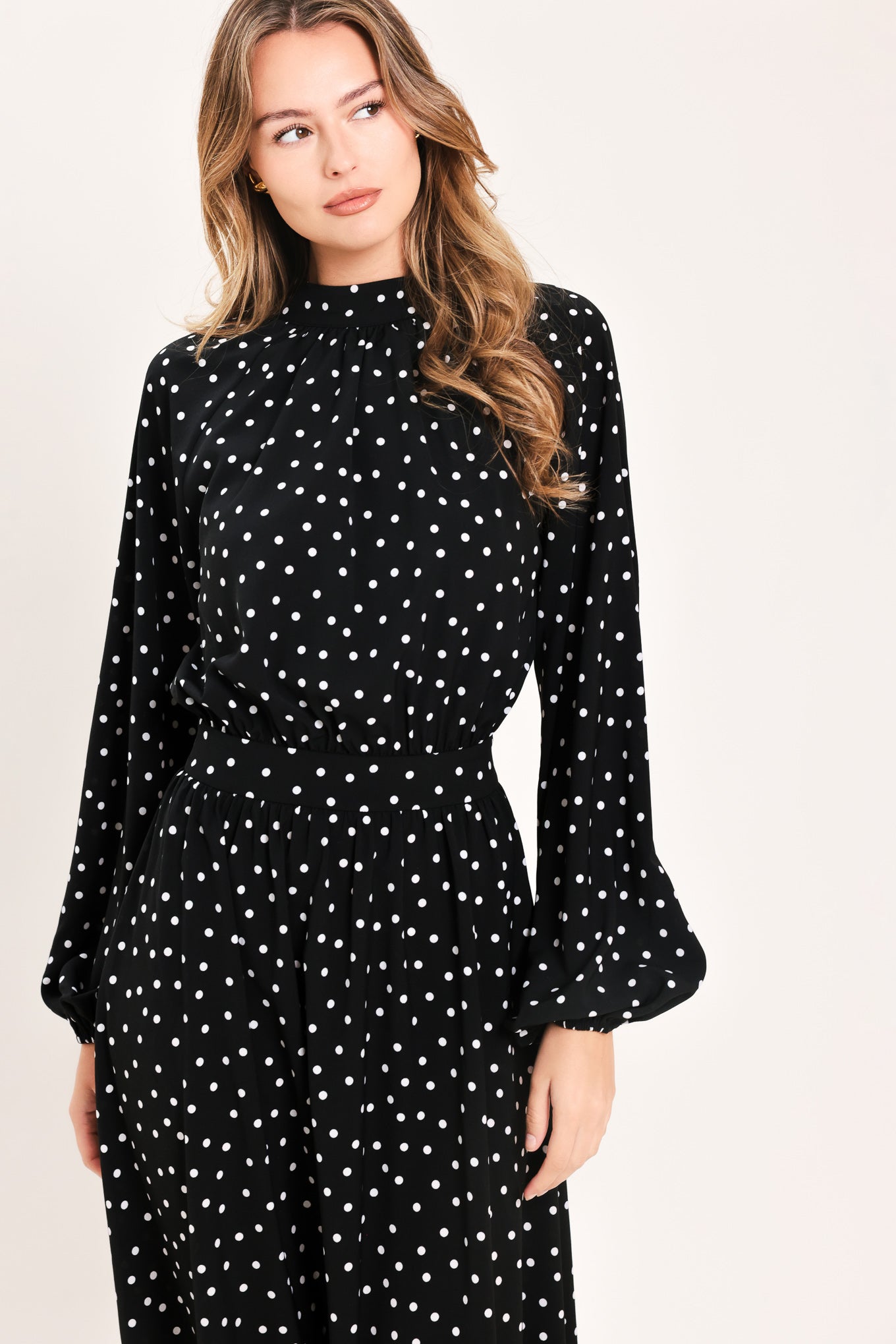 A woman modeling a black midi dress with a white polka dot pattern, featuring long balloon sleeves and a high mock neck | Flying Tomato