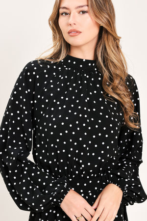 A woman modeling a black midi dress with a white polka dot pattern, featuring long balloon sleeves and a high mock neck | Flying Tomato