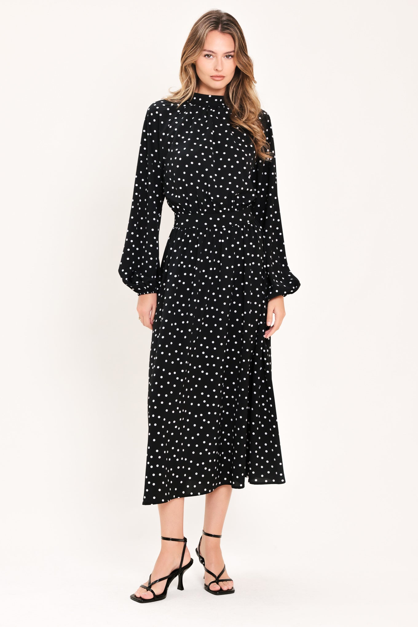 A woman modeling a black midi dress with a white polka dot pattern, featuring long balloon sleeves and a high mock neck | Flying Tomato