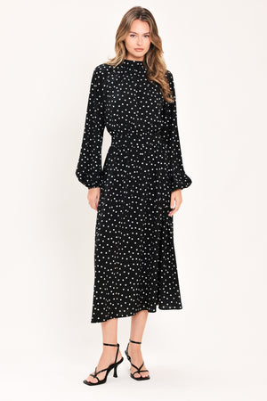 A woman modeling a black midi dress with a white polka dot pattern, featuring long balloon sleeves and a high mock neck | Flying Tomato