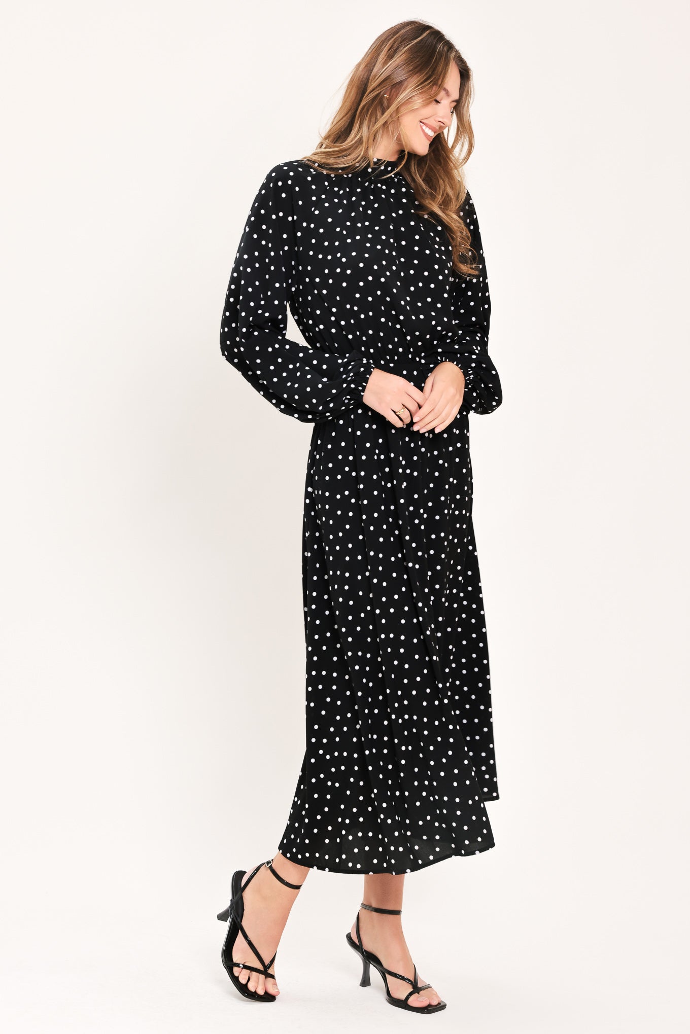 A woman modeling a black midi dress with a white polka dot pattern, featuring long balloon sleeves and a high mock neck | Flying Tomato