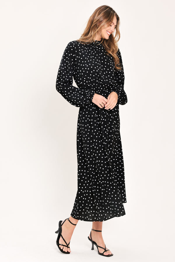 A woman modeling a black midi dress with a white polka dot pattern, featuring long balloon sleeves and a high mock neck | Flying Tomato