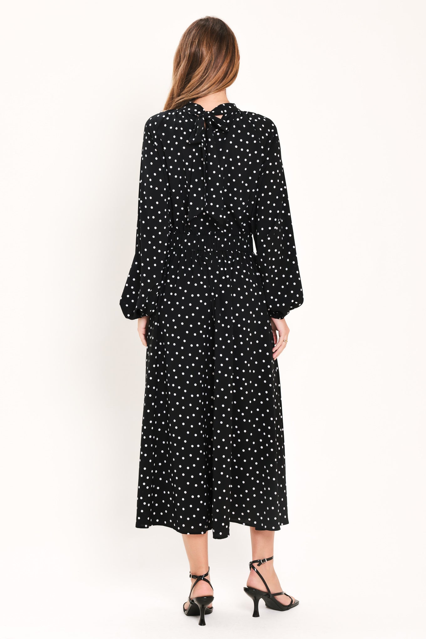 A woman modeling a black midi dress with a white polka dot pattern, featuring long balloon sleeves and a high mock neck | Flying Tomato