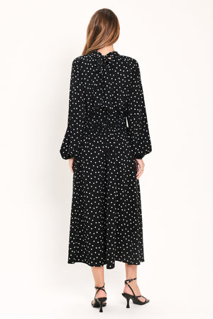 A woman modeling a black midi dress with a white polka dot pattern, featuring long balloon sleeves and a high mock neck | Flying Tomato