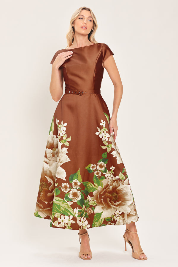 A woman models a rust satin midi dress with a floral print on the skirt, featuring white flowers, green leaves, and a matching brown belt. The dress also has cap sleeves and boatneck | Flying Tomato
