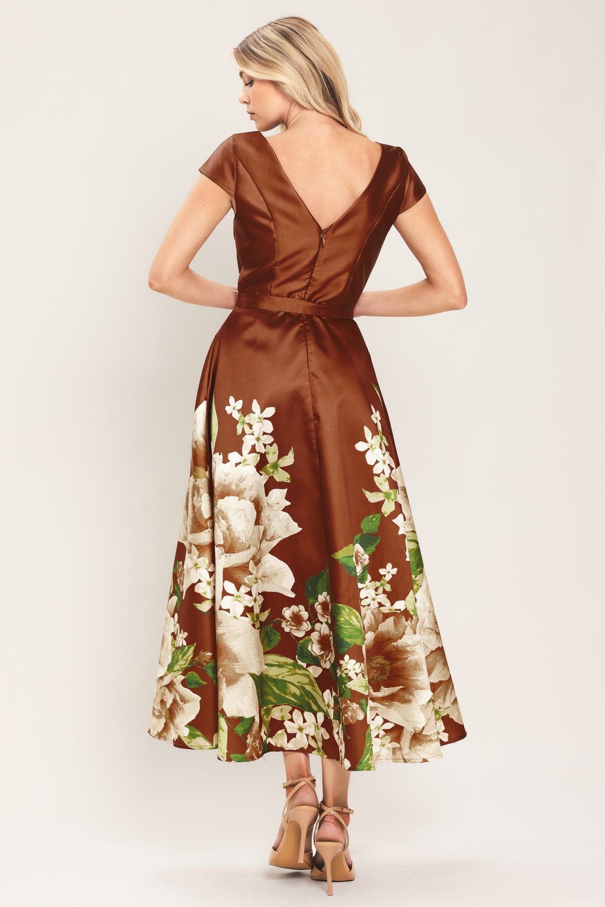 A woman models a rust satin midi dress with a floral print on the skirt, featuring white flowers, green leaves, and a matching brown belt. The dress also has cap sleeves and boatneck | Flying Tomato
