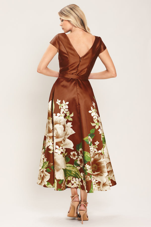 A woman models a rust satin midi dress with a floral print on the skirt, featuring white flowers, green leaves, and a matching brown belt. The dress also has cap sleeves and boatneck | Flying Tomato