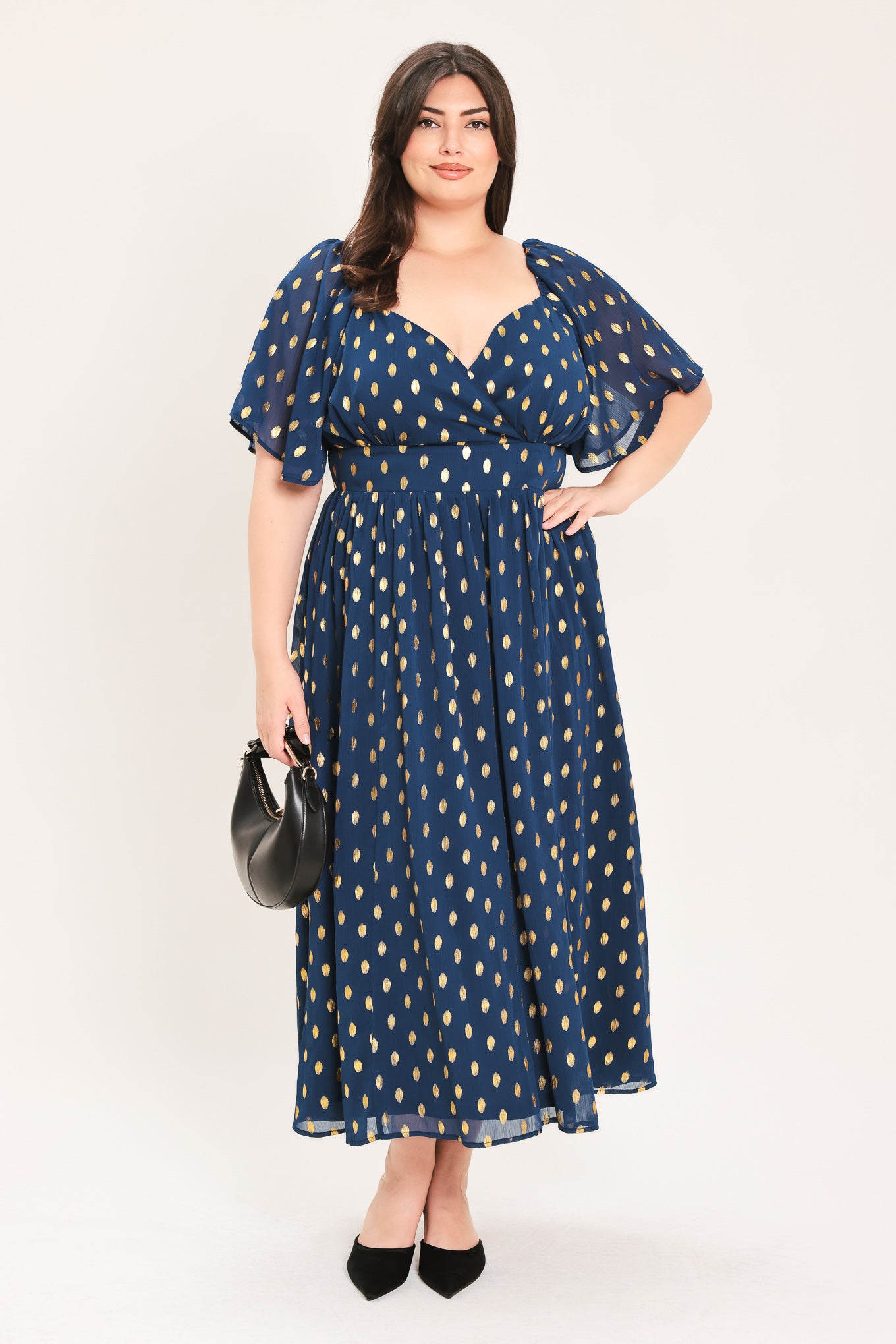 A woman modeling a light yellow midi dress with a small gold dotted pattern, featuring flutter sleeves and a V-neckline | Flying Tomato Curve