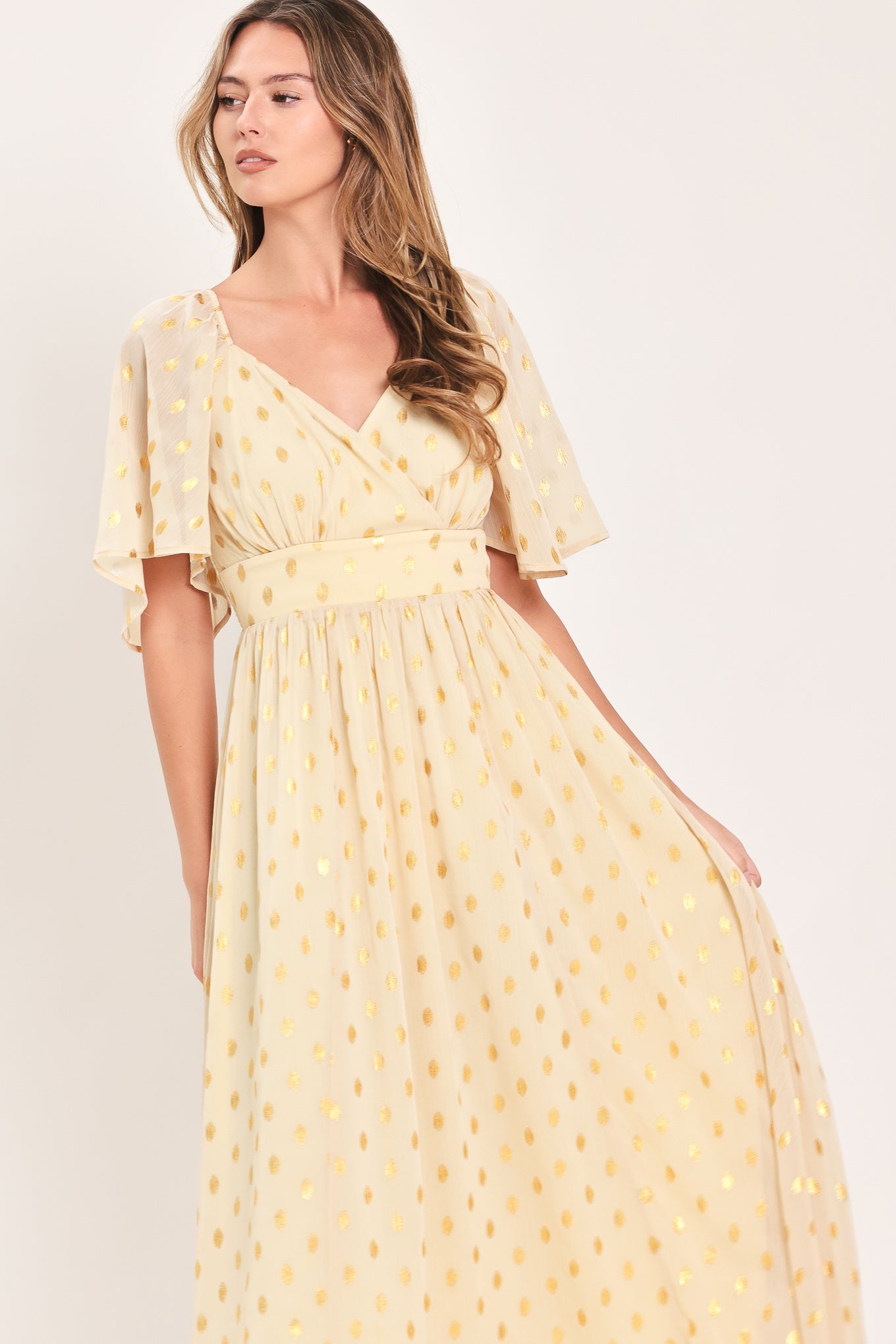 A woman modeling a light yellow midi dress with a small gold dotted pattern, featuring flutter sleeves and a V-neckline | Flying Tomato