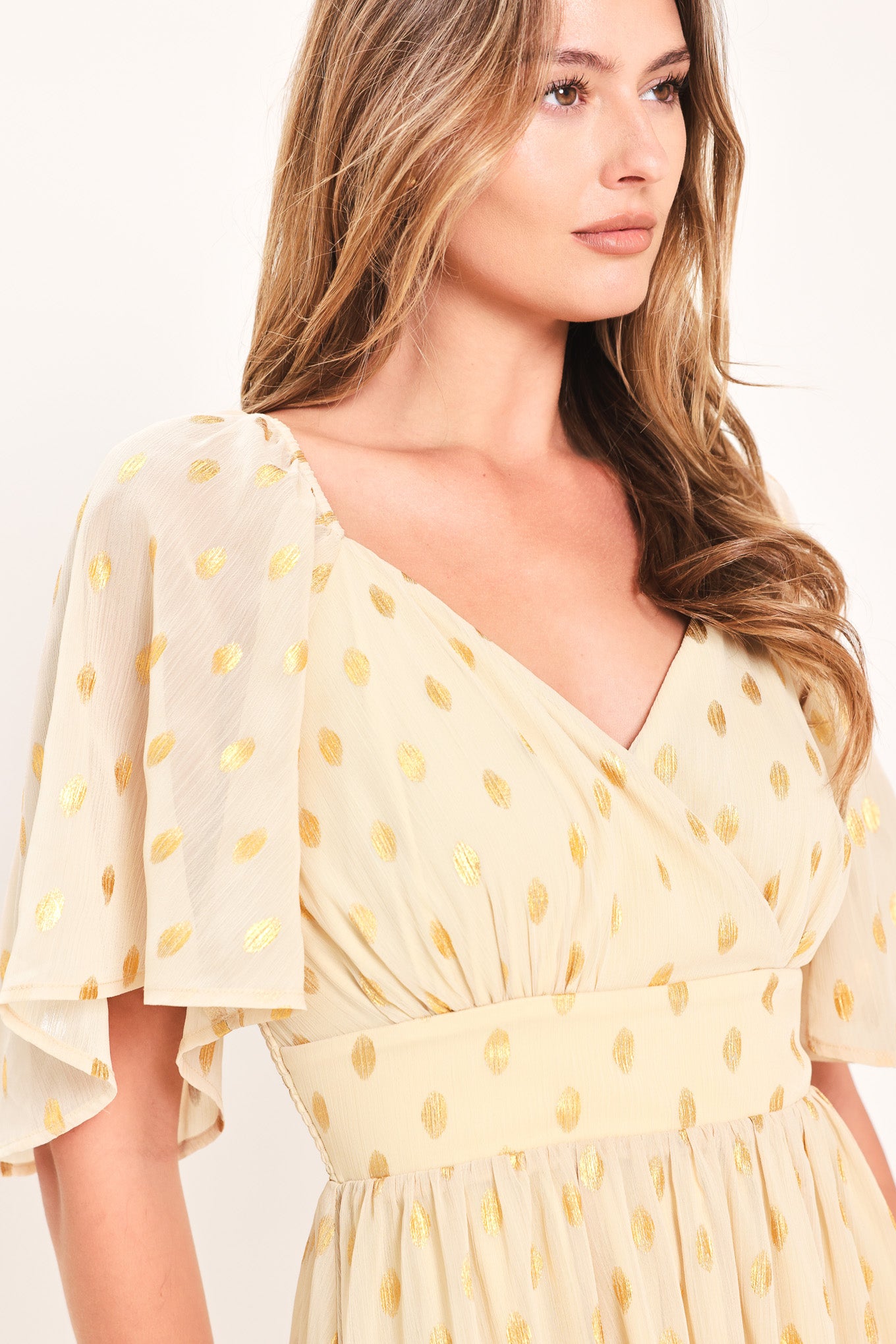 A woman modeling a light yellow midi dress with a small gold dotted pattern, featuring flutter sleeves and a V-neckline | Flying Tomato