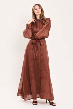 A woman wearing a rust-brown maxi dress with sheer balloon sleeves, a high ruffled collar, and a matching fabric tie belt at the waist | Flying Tomato