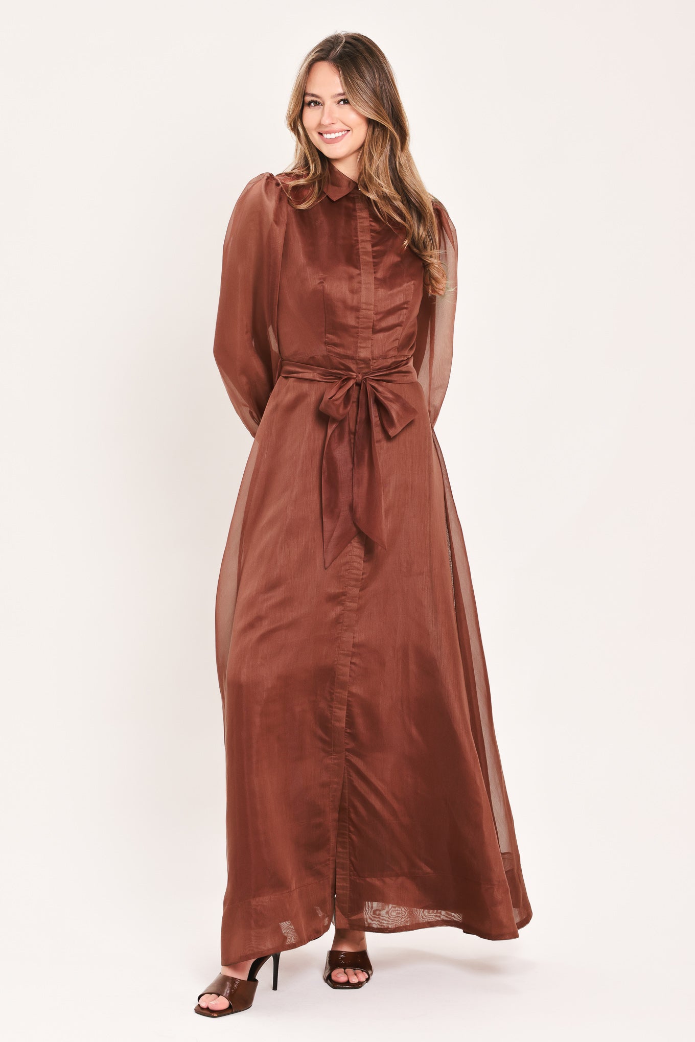 A woman wearing a rust-brown maxi dress with sheer balloon sleeves, a high ruffled collar, and a matching fabric tie belt at the waist | Flying Tomato