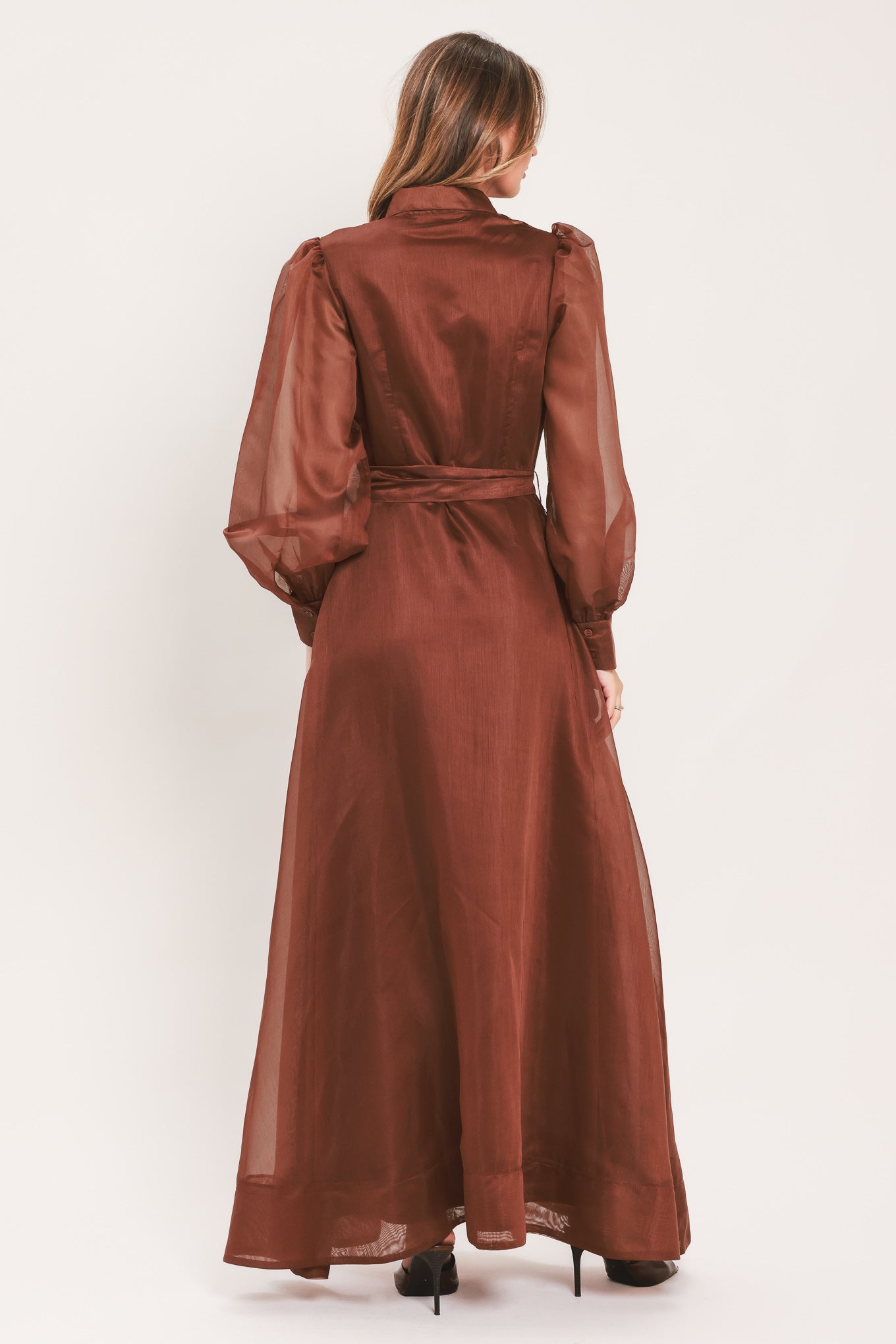 A woman wearing a rust-brown maxi dress with sheer balloon sleeves, a high ruffled collar, and a matching fabric tie belt at the waist | Flying Tomato