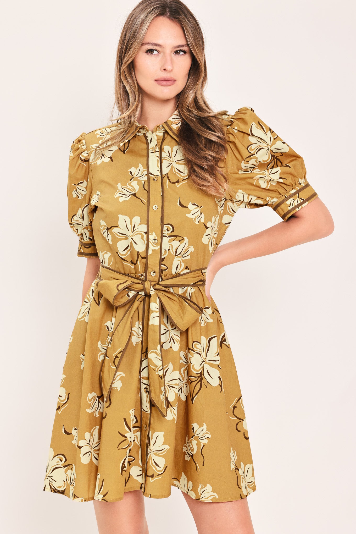 A woman wearing a camel yellow short-sleeve button-up dress with a beige and white floral print, featuring a tie waist belt | Flying Tomato