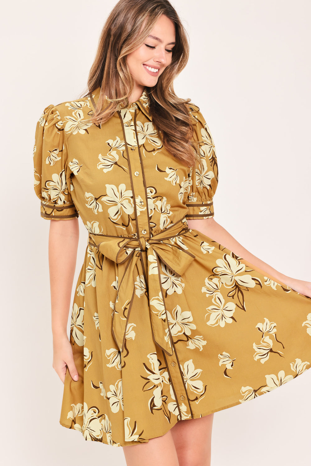 A woman wearing a camel yellow short-sleeve button-up dress with a beige and white floral print, featuring a tie waist belt | Flying Tomato