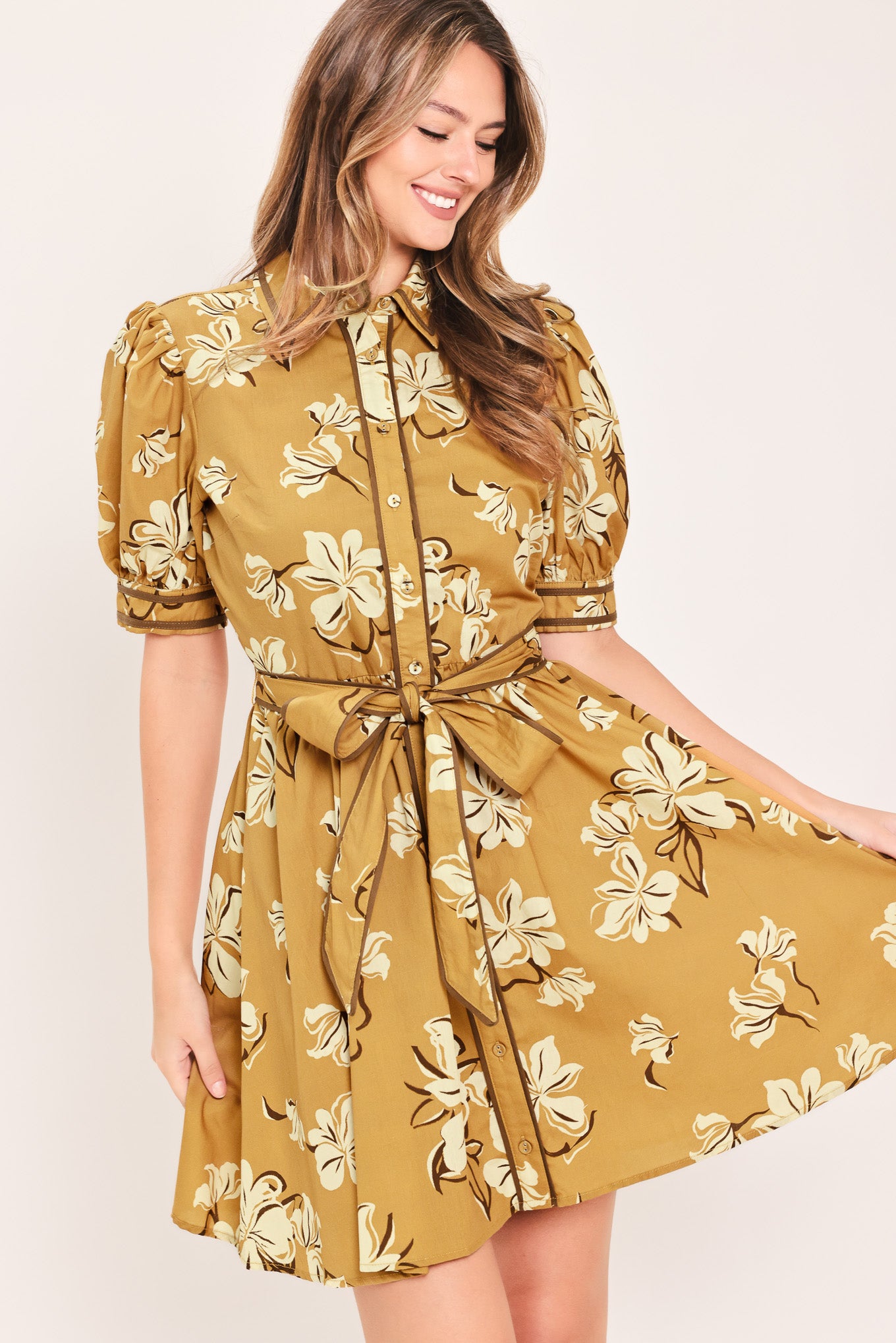 A woman wearing a camel yellow short-sleeve button-up dress with a beige and white floral print, featuring a tie waist belt | Flying Tomato