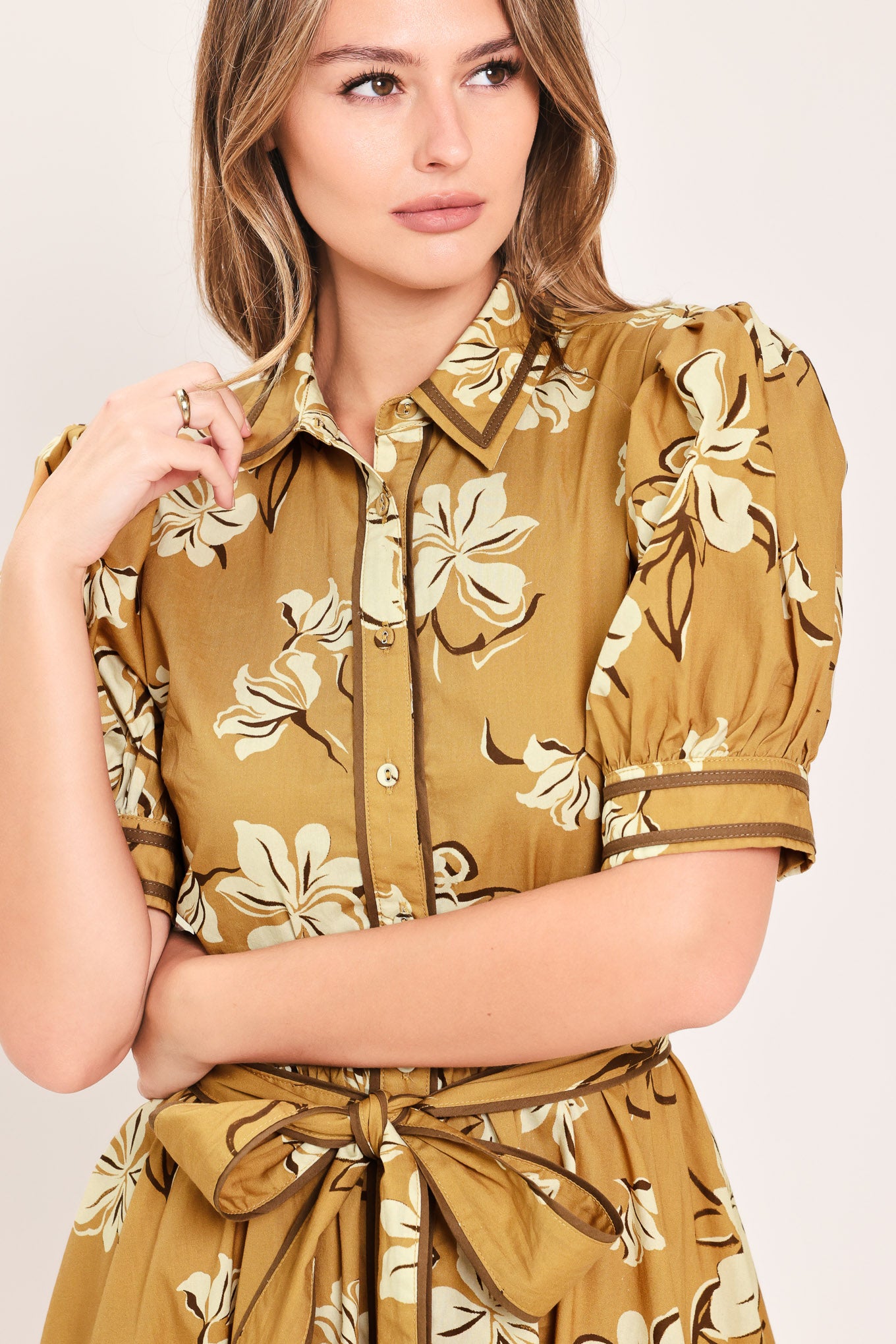 A woman wearing a camel yellow short-sleeve button-up dress with a beige and white floral print, featuring a tie waist belt | Flying Tomato