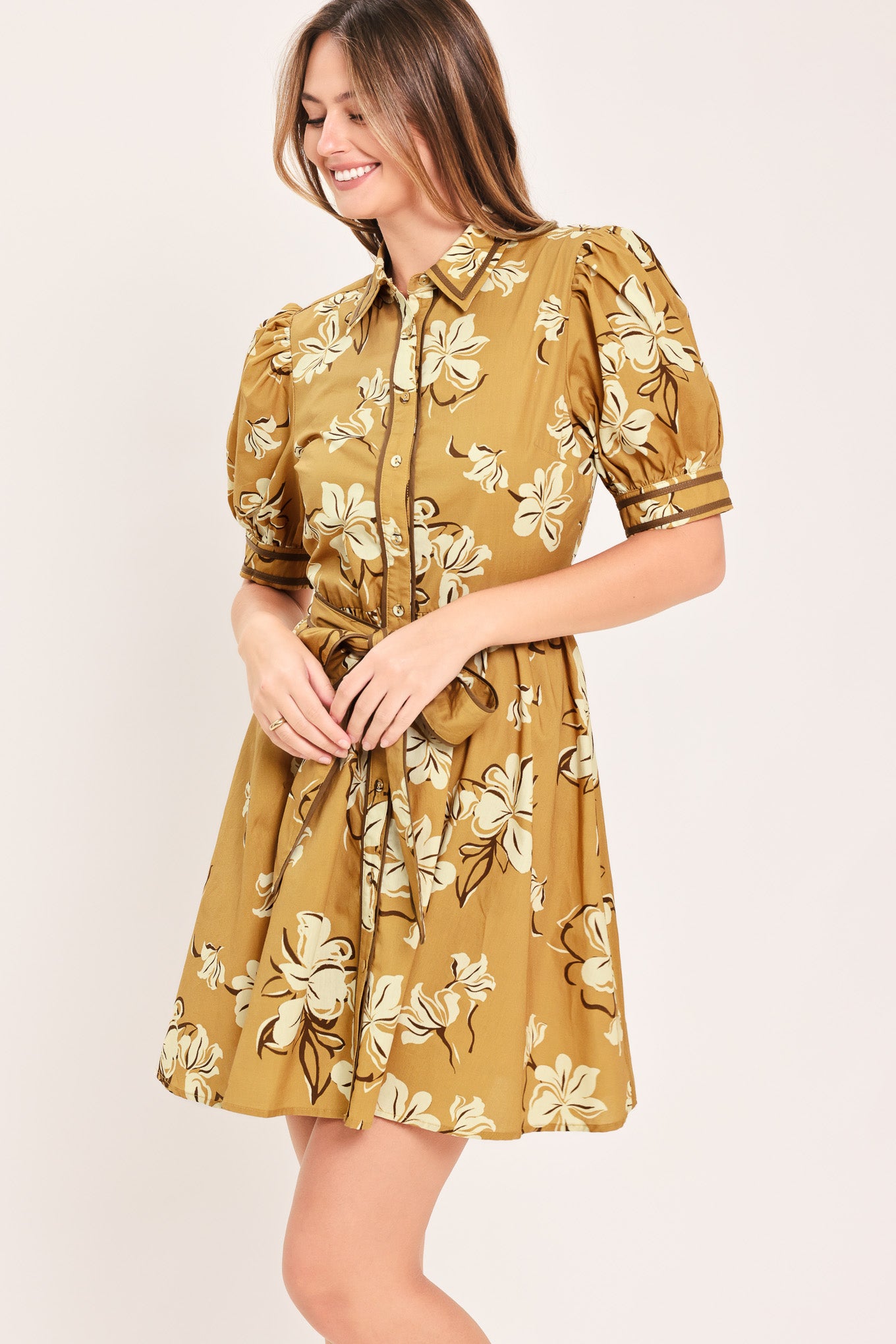 A woman wearing a camel yellow short-sleeve button-up dress with a beige and white floral print, featuring a tie waist belt | Flying Tomato