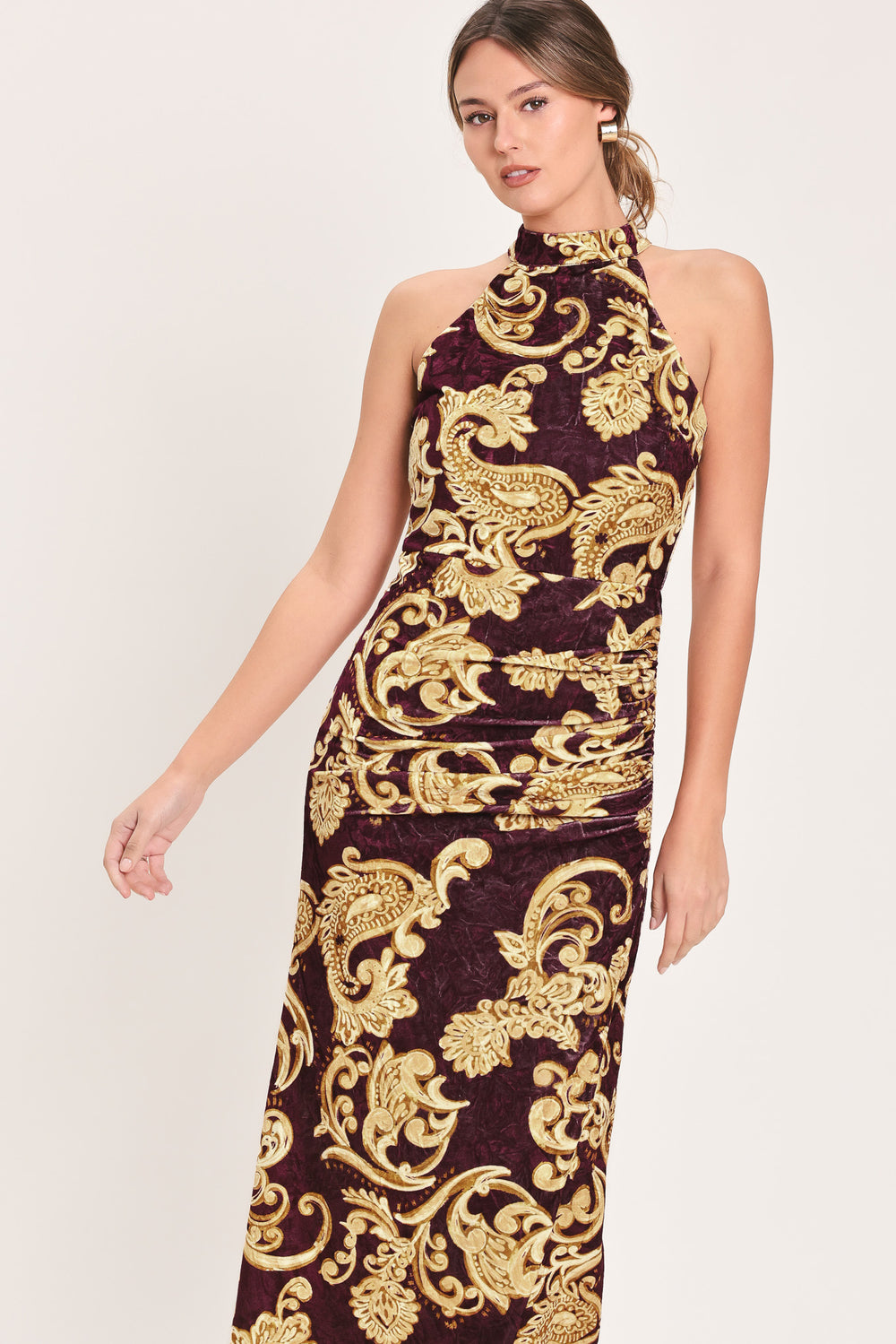 Woman wearing a mock halter neck burgundy velvet midi dress with side ruching, front side leg opening, and gold paisley baroque print | Flying Tomato