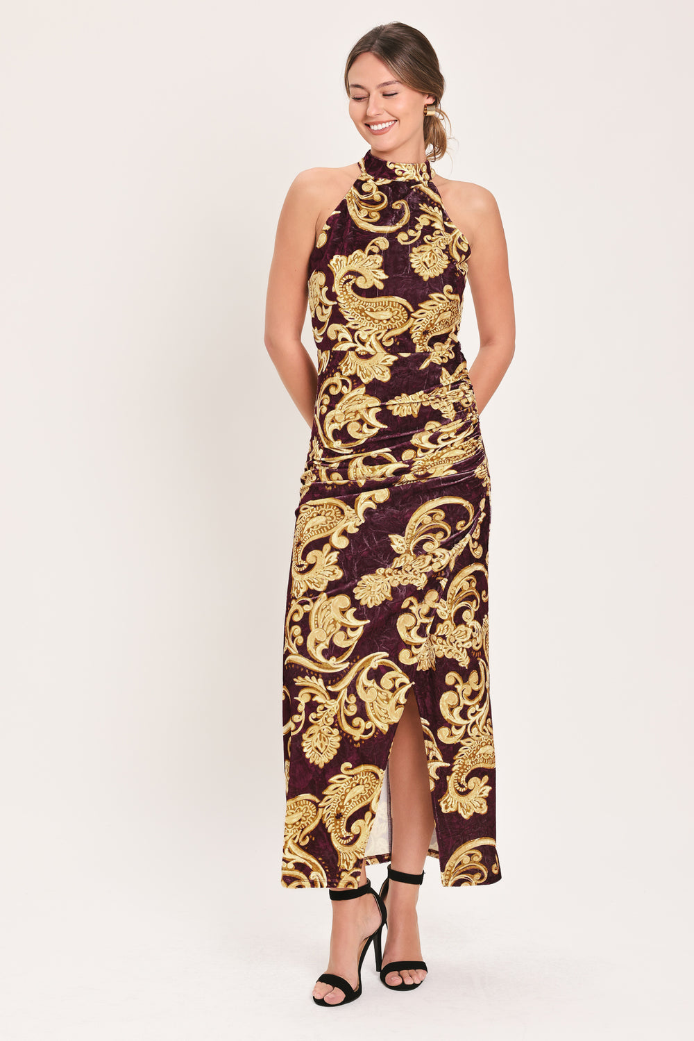 Woman wearing a mock halter neck burgundy velvet midi dress with side ruching, front side leg opening, and gold paisley baroque print | Flying Tomato
