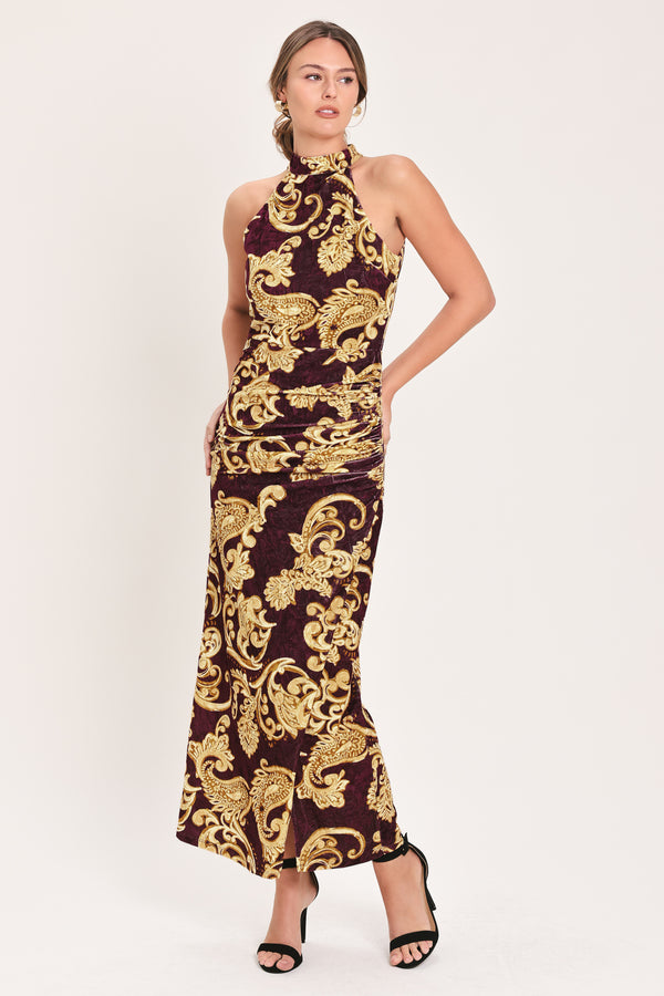 Woman wearing a mock halter neck burgundy velvet midi dress with side ruching, front side leg opening, and gold paisley baroque print | Flying Tomato