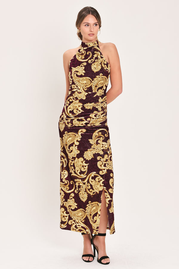 Woman wearing a mock halter neck burgundy velvet midi dress with side ruching, front side leg opening, and gold paisley baroque print | Flying Tomato