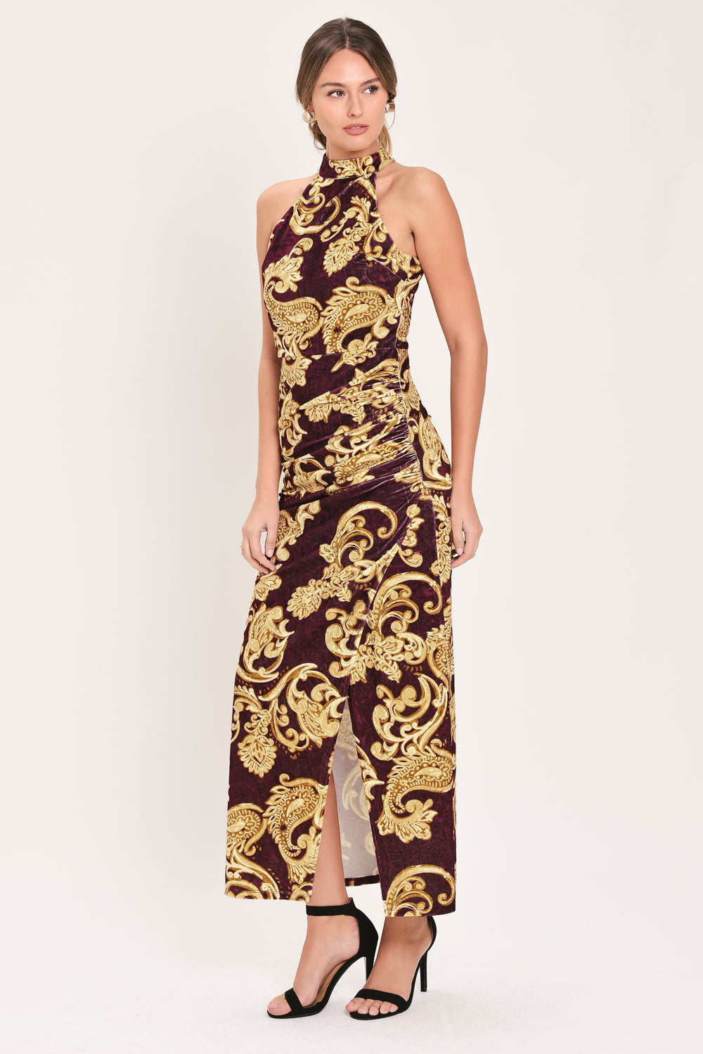 Woman wearing a mock halter neck burgundy velvet midi dress with side ruching, front side leg opening, and gold paisley baroque print | Flying Tomato