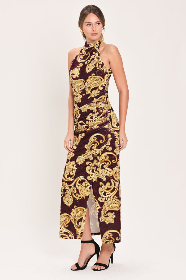 Woman wearing a mock halter neck burgundy velvet midi dress with side ruching, front side leg opening, and gold paisley baroque print | Flying Tomato