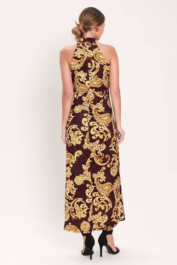 Woman wearing a mock halter neck burgundy velvet midi dress with side ruching, front side leg opening, and gold paisley baroque print | Flying Tomato