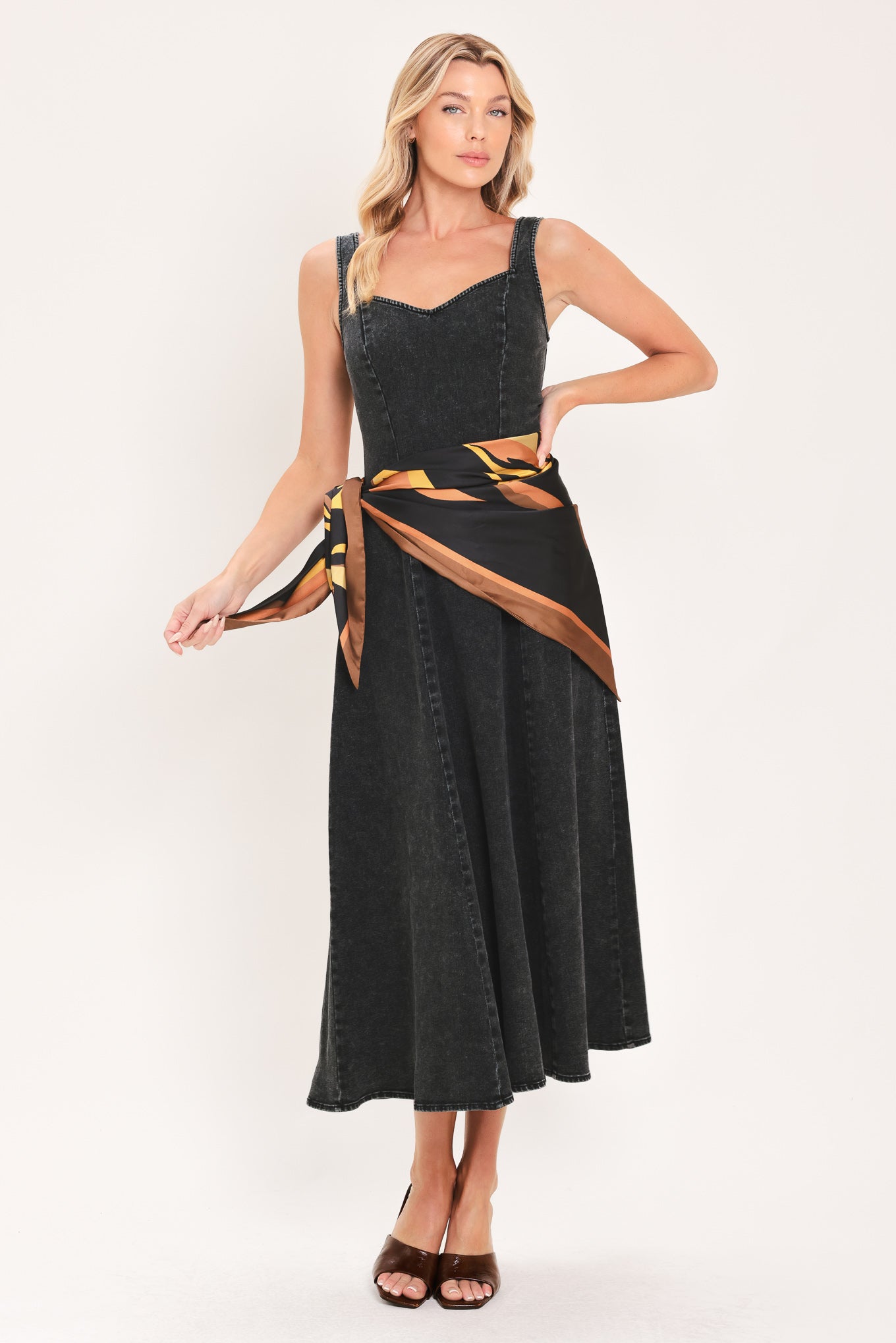 A woman modeling a sleeveless black denim midi dress with a scarf wrap-style belt featuring a brown and gold geometric pattern | Flying Tomato