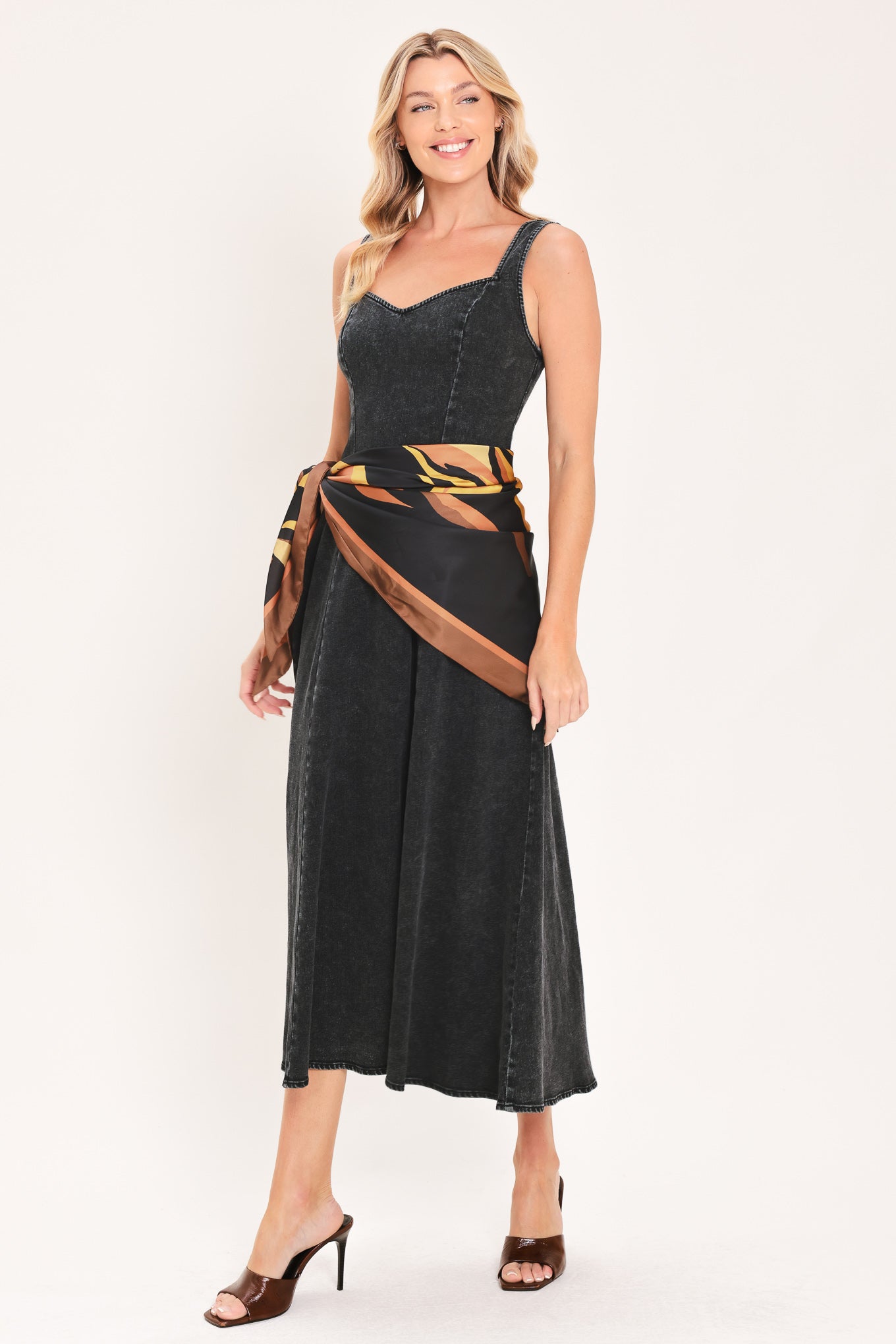 A woman modeling a sleeveless black denim midi dress with a scarf wrap-style belt featuring a brown and gold geometric pattern | Flying Tomato