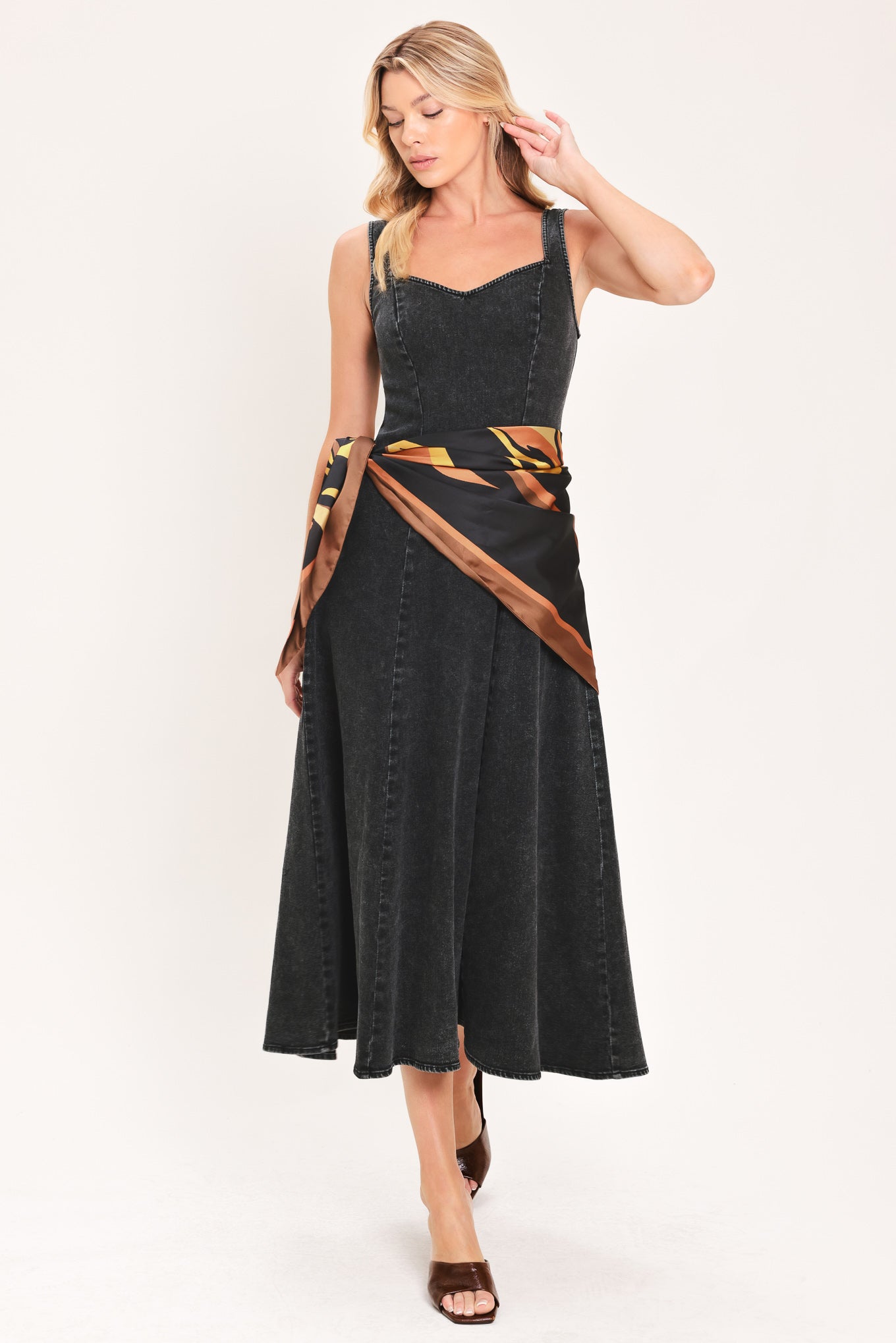 A woman modeling a sleeveless black denim midi dress with a scarf wrap-style belt featuring a brown and gold geometric pattern | Flying Tomato