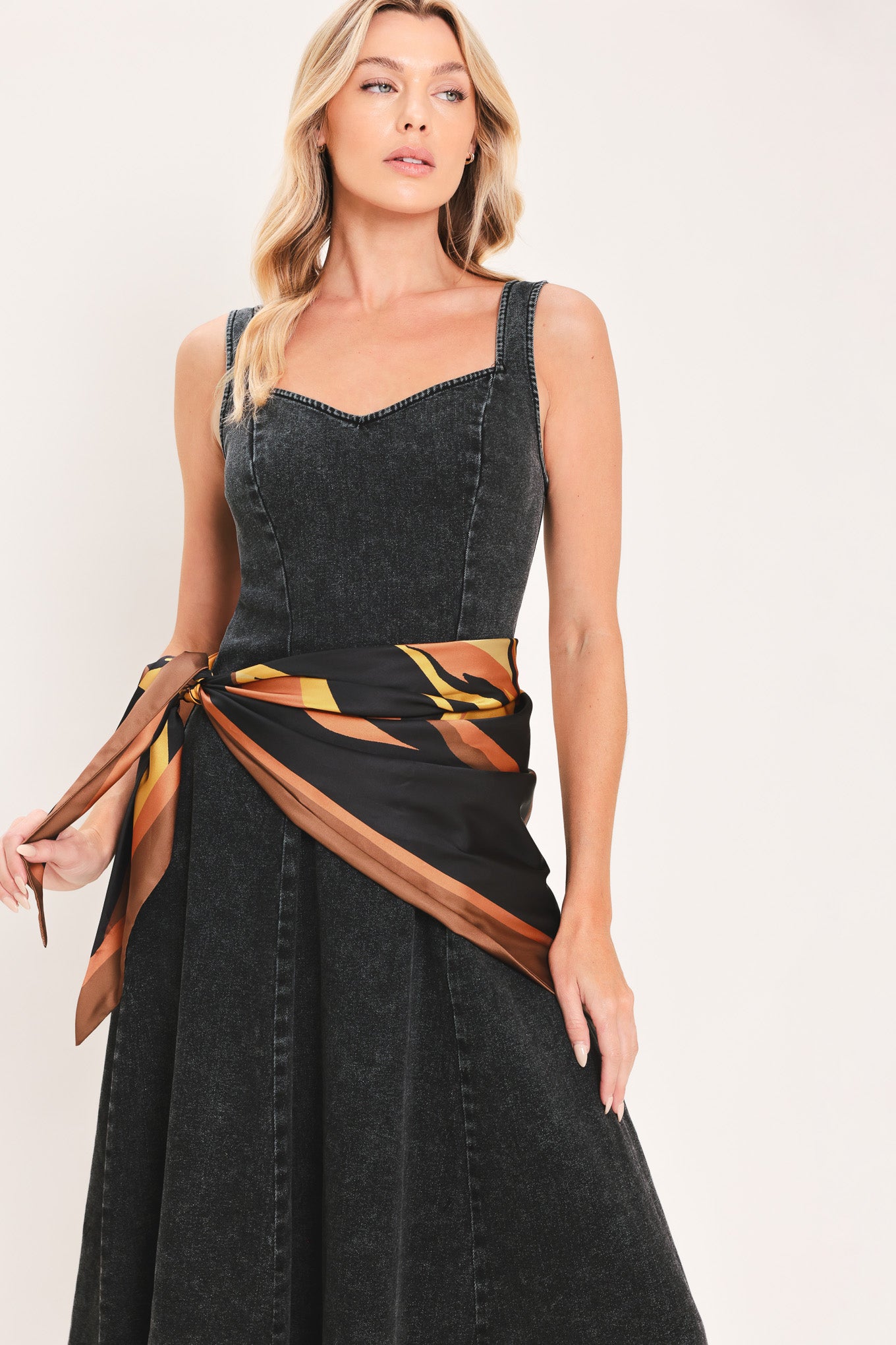 A woman modeling a sleeveless black denim midi dress with a scarf wrap-style belt featuring a brown and gold geometric pattern | Flying Tomato