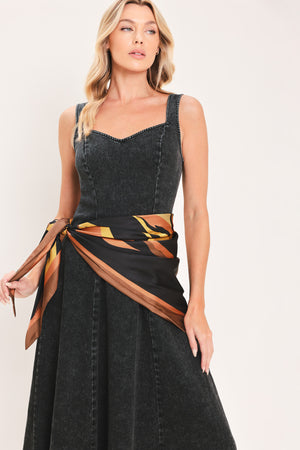 A woman modeling a sleeveless black denim midi dress with a scarf wrap-style belt featuring a brown and gold geometric pattern | Flying Tomato