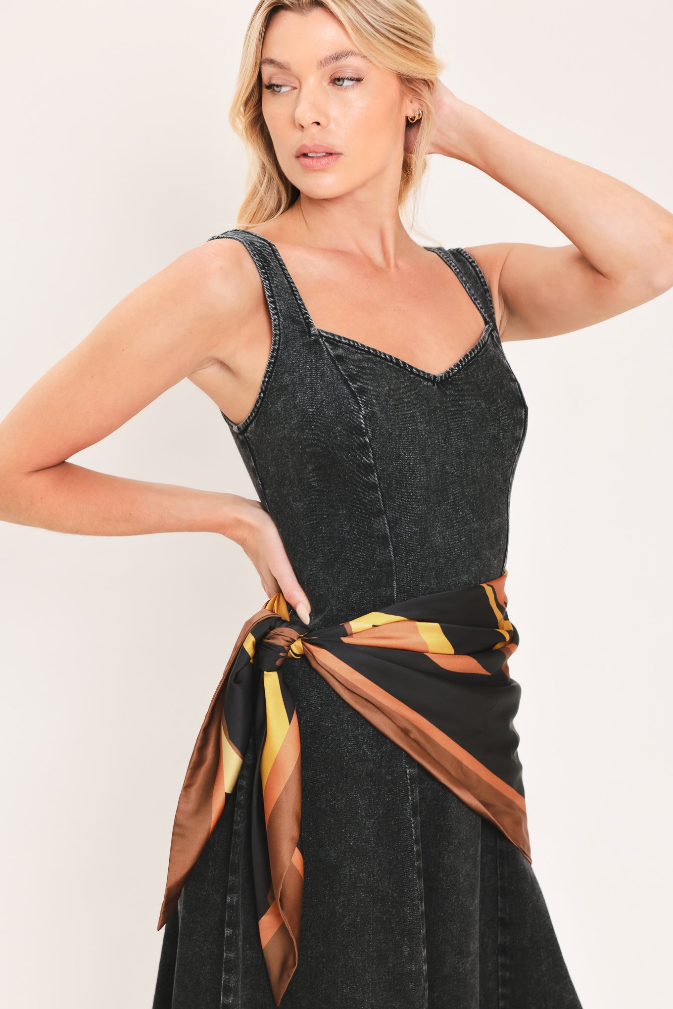 A woman modeling a sleeveless black denim midi dress with a scarf wrap-style belt featuring a brown and gold geometric pattern | Flying Tomato