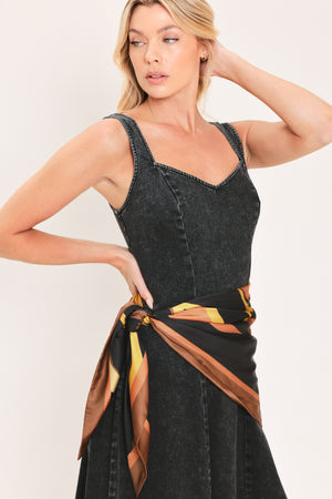 A woman modeling a sleeveless black denim midi dress with a scarf wrap-style belt featuring a brown and gold geometric pattern | Flying Tomato