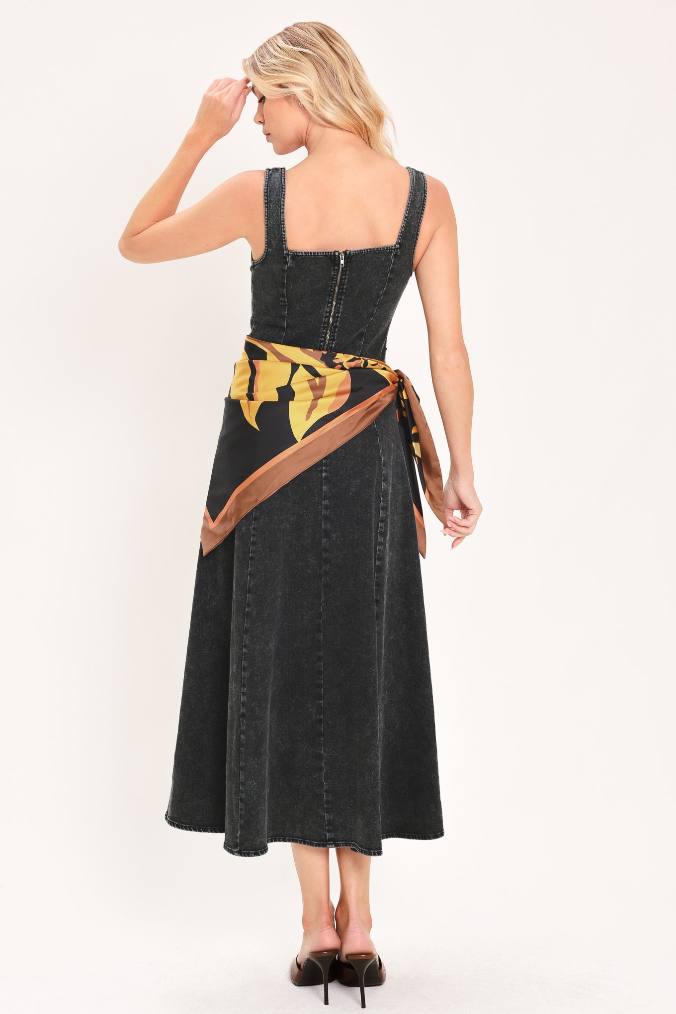 A woman modeling a sleeveless black denim midi dress with a scarf wrap-style belt featuring a brown and gold geometric pattern | Flying Tomato
