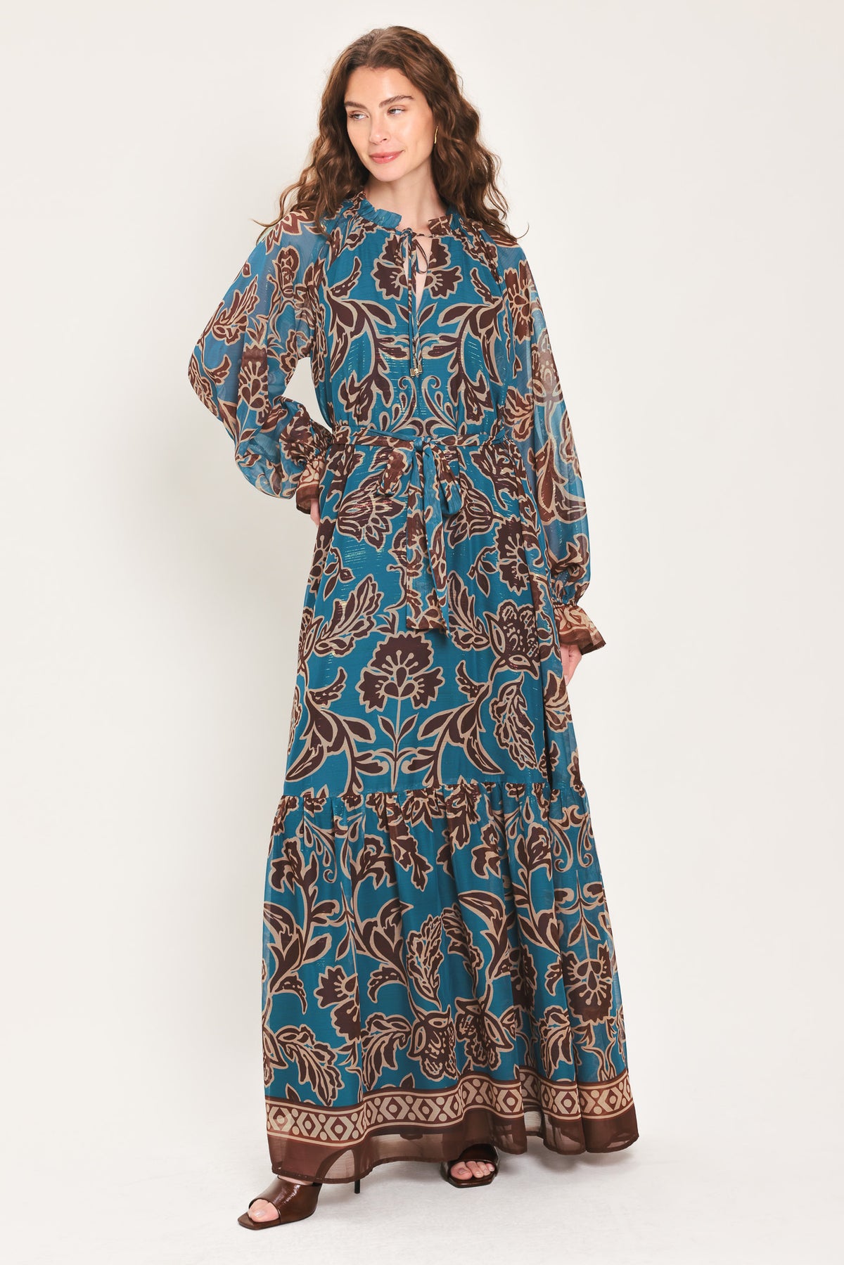 Woman wearing a teal blue long sleeve maxi dress with ruffled sleeves, tiered skirts, and brown floral print and neck tie | Flying Tomato