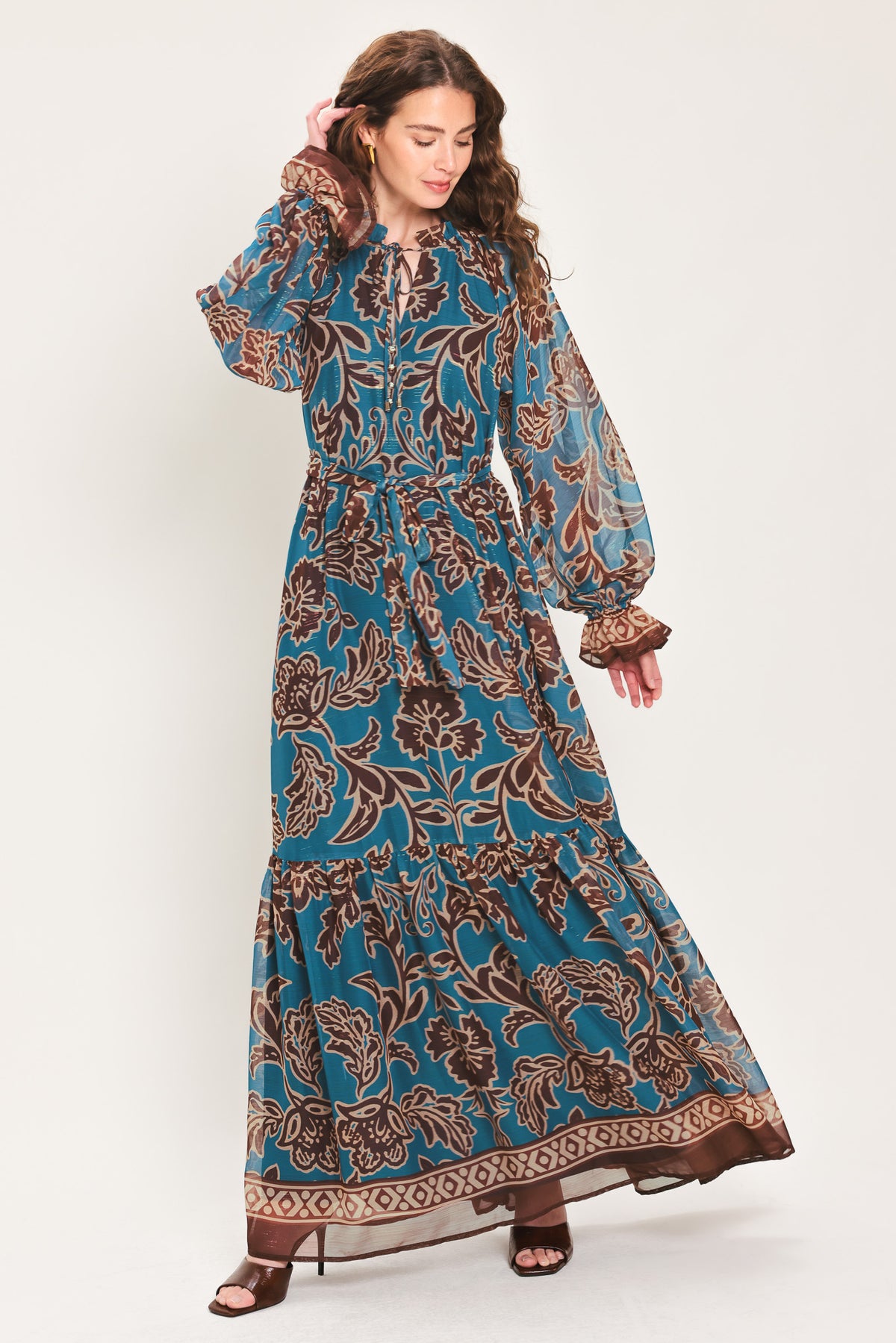 Woman wearing a teal blue long sleeve maxi dress with ruffled sleeves, tiered skirts, and brown floral print and neck tie | Flying Tomato