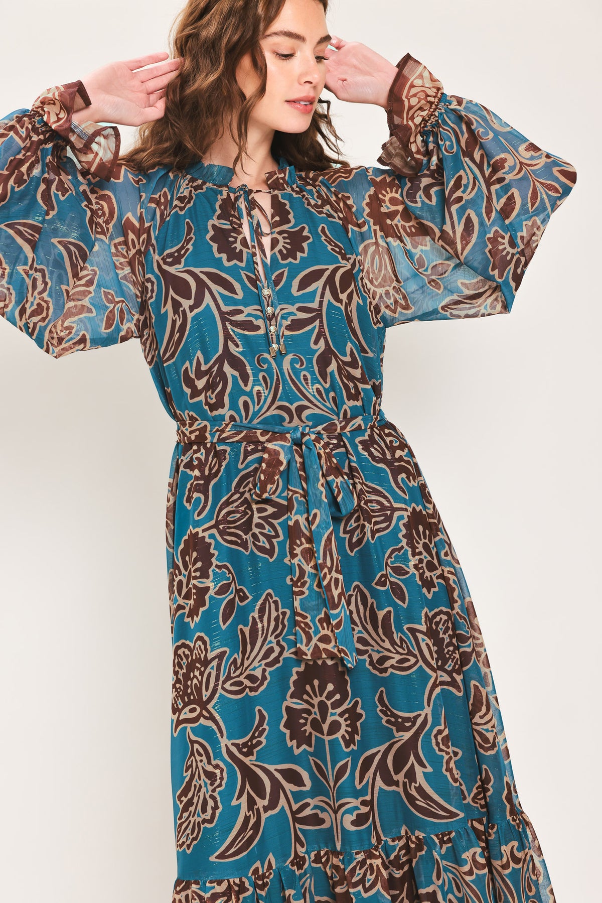 Woman wearing a teal blue long sleeve maxi dress with ruffled sleeves, tiered skirts, and brown floral print and neck tie | Flying Tomato